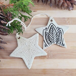 May include: Two off-white ceramic star ornaments with twine hangers. One star has a snowflake design, the other a black leaf pattern. The ornaments are on a wooden surface with greenery and pinecones.
