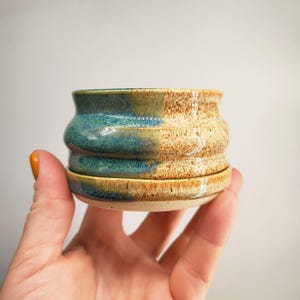 May include: A handmade ceramic plant pot with a matching saucer. The pot has a two-tone glaze, with teal and brown sections. The rounded body has a textured, ribbed design. The saucer is the same colour and shape as the pot.