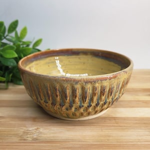 May include: A handmade ceramic bowl with a textured exterior. The bowl has a yellow interior and a brown and blue exterior with a textured pattern. The bowl is on a wooden surface.