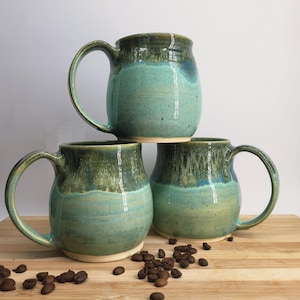 May include: Three ceramic mugs stacked in a pyramid formation. Each mug features a rounded body with a handle and a two-tone glaze, transitioning from a deep forest green at the rim to a lighter, speckled turquoise blue on the lower half.