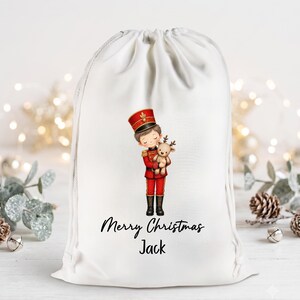 May include: A white drawstring gift bag featuring a printed illustration of a child dressed as a toy soldier in a red uniform holding a small reindeer plush toy. Below the illustration, the text reads Merry Christmas Jack in black script.