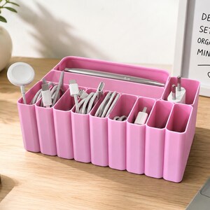 Könnte beinhalten: Ein rosa Schreibtisch-Organizer aus Kunststoff mit mehreren vertikalen Fächern, die verschiedene weiße Ladekabel, einen weißen Netzstecker und einen silbernen Stift enthalten. Der Organizer hat eine rechteckige Form mit abgerundeten Kanten.