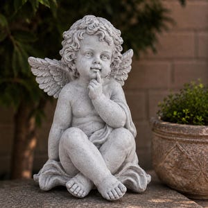 May include: A light gray stone-colored statue of a cherub with curly hair and feathered wings. The figure is seated with legs crossed, holding one index finger to its lips in a quiet gesture. The sculpture features detailed facial expressions.