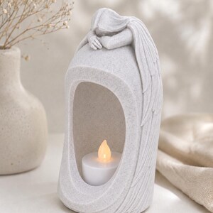 May include: A light gray, speckled stone-effect candle holder shaped like a mourning angel with wings draped down the sides. The figure rests its head on its arms atop a hollow archway containing a small, white, battery-operated tea light candle.