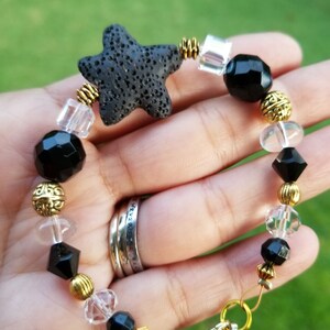 May include: A beaded bracelet featuring a black lava stone starfish, faceted black beads, clear crystal beads, and gold-colored metal accents. The bracelet is strung on a flexible wire and has a gold-colored clasp.