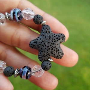 May include: A handmade bracelet featuring a black lava stone starfish charm. The bracelet includes clear and purple glass beads, and small black lava stone beads. Silver-toned metal accents add detail to this unique jewelry piece.