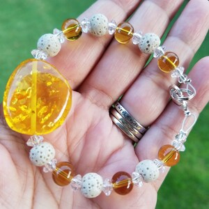 May include: A handcrafted bracelet featuring a large, polished amber stone and a mix of round, amber-colored beads, clear crystal accents, and textured, off-white beads. The bracelet is secured with a silver-colored clasp.