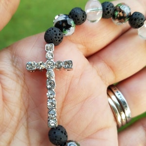 May include: A beaded bracelet featuring a silver cross adorned with clear crystals. The bracelet incorporates black lava beads and decorative glass beads with floral designs. The bracelet is held in a hand against a blurred green background.
