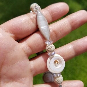 May include: A handmade bracelet featuring a variety of natural elements. The bracelet includes a large, elongated gray bead, small round porous beads, and a spiral seashell. Clear and purple beads add accents. The clasp is silver.