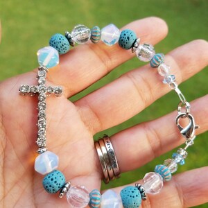 May include: A beaded bracelet featuring a silver cross pendant with clear rhinestones. The bracelet includes blue lava rock beads, clear faceted beads, and iridescent white beads. The bracelet is secured with a silver clasp.