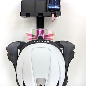 Peut inclure: Un support de rangement mural noir portant un casque blanc avec la marque Oakley, une paire de lunettes de soleil KOO aux verres teintés rose et un compteur de vélo Hammerhead noir affichant 86 % avec un câble de charge rose branché.