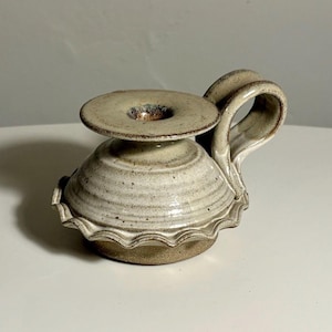 May include: A ceramic candle holder with a speckled off-white glaze and a brown base. The piece features a wide, flat top with a central opening, a rounded body with horizontal ridges, a scalloped bottom edge, and a curved handle on the side.