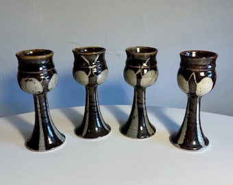 May include: Four ceramic goblets with a tall, slender stem and a flared base. Each goblet features a dark brown glaze with contrasting cream-coloured, rounded vertical patterns that extend from the bowl down to the base of the stem.