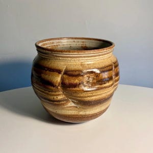May include: A round ceramic pot featuring a variegated glaze of tan, beige, and dark brown horizontal bands. The vessel has a wide opening with a defined rim and a small, square-shaped stamped mark on the side, showcasing a textured finish.