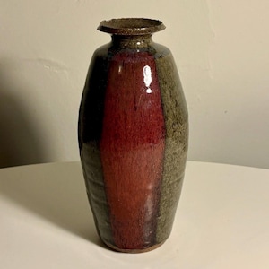 May include: A ceramic vase with a cylindrical body and a narrow neck featuring a flared rim. The surface displays a vertical color block design with a deep red center panel flanked by textured olive green sides and a dark brown glazed rim.