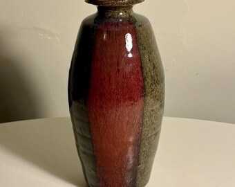 May include: A ceramic vase with a cylindrical body and a narrow neck featuring a flared rim. The surface displays a vertical colour block design with a deep red centre panel flanked by textured olive green sides and a dark brown glazed rim.
