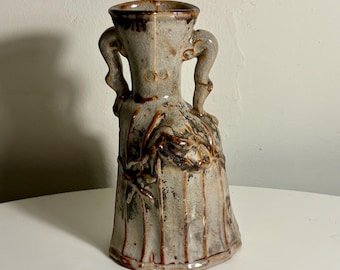 May include: A ceramic vase featuring a light grey glaze with brown vertical streaks and a crackled texture. The vessel has two curved handles on the sides, a wide flared rim, and a tapered body with a moulded relief design of branches.