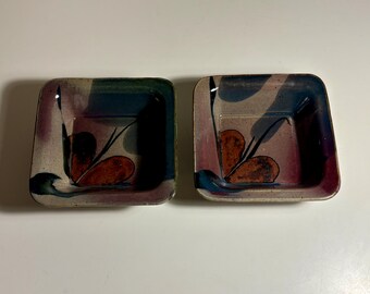 May include: Two square ceramic bowls featuring a speckled beige base with hand-painted abstract floral motifs in shades of deep purple, dark blue, and earthy brown. The bowls have rounded corners and a smooth, glazed finish throughout.