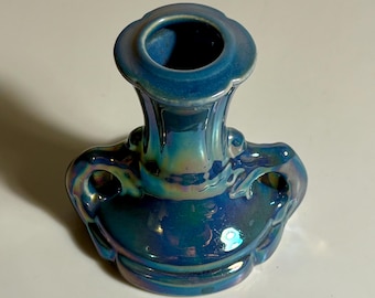 May include: A ceramic candle holder with a glossy, iridescent blue finish. The object features a central cylindrical opening for a candle, a wide circular base, and two curved handles on opposite sides, reflecting light with a metallic sheen.