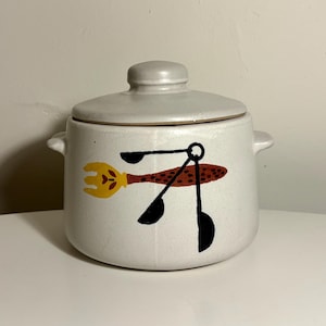 Peut inclure: Un pot en céramique blanc cassé moucheté avec un couvercle assorti et deux poignées latérales. L'avant présente une illustration graphique d'une fourchette jaune et de trois cuillères à mesurer noires avec un motif de manche texturé rouille.