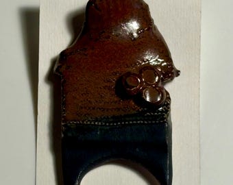 May include: A ceramic pin featuring a glossy brown upper section with three small raised circular accents and a matte dark blue lower section with two pointed legs. The word PIN is printed in black ink at the bottom of the display card.
