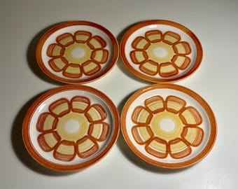May include: A set of four round ceramic plates featuring a central cream-coloured circle surrounded by a ring of eight stylised orange and yellow petals. Each plate has a wide, solid orange rim and a smooth, glossy finish throughout.