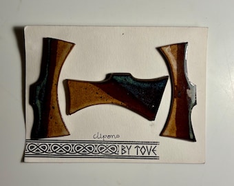 May include: Three ceramic hair clips with a glossy finish in shades of brown, tan, and teal. The clips are mounted on a white card featuring the text clipono and BY TOVE with a decorative border pattern at the bottom of the display card.