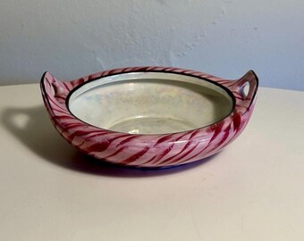 May include: A shallow glass bowl featuring a swirling pattern of deep pink and white stripes. The interior has an iridescent, pearlescent finish, while the exterior rim is accented with a thin black line. It includes two small, integrated handles.
