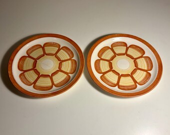 May include: Two round ceramic plates featuring a white centre and a wide orange rim. The surface of each plate is decorated with a repeating pattern of eight stylised orange and yellow petals arranged in a circular floral design.