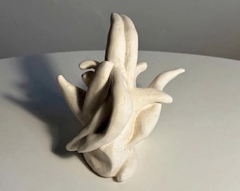 May include: A sculptural object made of off-white ceramic with a matte finish. The piece features multiple elongated, curved, and pointed protrusions extending from a central base, creating an abstract, organic form with subtle speckled texture.