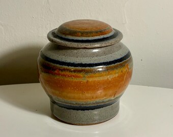 May include: A ceramic jar with a matching lid, featuring a rounded body and a speckled grey base. The surface is decorated with horizontal bands of deep blue, burnt orange, and tan glaze, creating a layered, multi-coloured appearance.