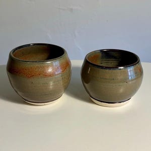 May include: Two rounded ceramic cups with a smooth, glossy finish. The glaze features a blend of earthy olive green and warm, burnt orange tones, with a dark rim and a clean, unglazed white base. The vessels have a wide, bulbous shape.