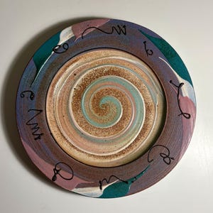 May include: A round ceramic plate featuring a central spiral pattern in shades of beige, light blue, and brown. The wide rim is decorated with patches of teal, purple, and white, accented by thin, dark brown, hand-drawn squiggly line motifs.