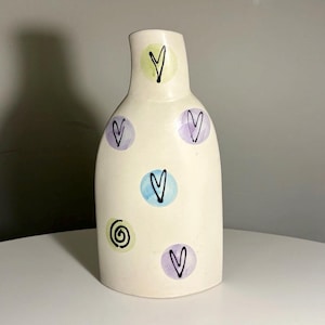 Peut inclure: Un vase en céramique de couleur crème avec un col étroit et un corps arrondi. La surface est décorée de cercles pastel peints à la main en vert clair, bleu et violet, chacun contenant un cœur dessiné à la main ou un motif en spirale.