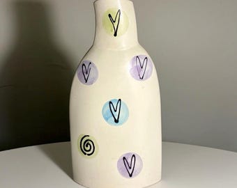 May include: A cream-coloured ceramic vase with a narrow neck and rounded body. The surface is decorated with hand-painted pastel circles in light green, blue, and purple, each containing a black hand-drawn heart or a spiral pattern design.