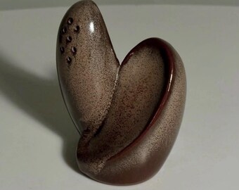 May include: A ceramic salt shaker with a curved, abstract form and a speckled brown and tan glaze. The top section features a cluster of small holes for dispensing salt, while the body has a smooth, rounded silhouette with a deep central groove.