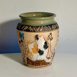 May include: A ceramic vase with a light tan base featuring a relief design of two cats sitting on a wooden log. One cat is white with black spots, and the other is white with orange patches. A blue butterfly and brown floral stems are visible.