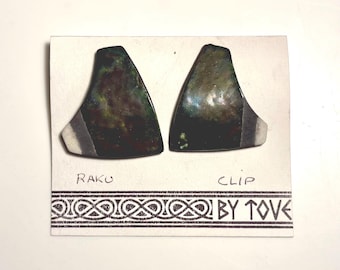 May include: A pair of asymmetrical clip-on earrings featuring a dark, iridescent finish with green and copper tones. The earrings are mounted on a white display card with the text RAKU CLIP BY TOVE and a decorative border pattern.