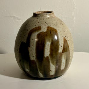 May include: A round ceramic vase with a speckled beige base and a glossy finish. The surface features vertical, abstract brushstrokes in dark brown and white, creating a layered, textured appearance. The vase has a narrow, circular opening at the top.