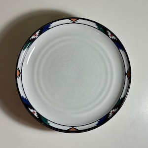 May include: A round white ceramic plate featuring a decorative border pattern. The rim is adorned with a repeating geometric design in shades of dark blue, green, red, and peach, enclosed by thin black lines that circle the edge of the plate.
