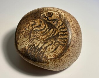 May include: A rounded, beige ceramic stone with a speckled texture and a dark brown, incised abstract pattern on its surface. The object has a smooth, polished appearance with a curved, irregular shape and a mottled, earthy colour palette.
