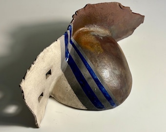 May include: A sculptural ceramic object with a rounded, brown-toned body and a flat, off-white side panel. The piece features two small square cutouts and is decorated with three distinct vertical stripes in deep blue and dark grey.