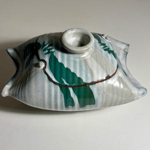 May include: A ceramic lidded vessel with a ribbed, off-white body featuring hand-painted green brushstroke patterns. The lid has a central circular handle and a dark brown rim, while the overall shape is elongated with pointed, flared ends.