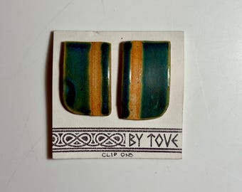 May include: A pair of rectangular ceramic clip-on earrings featuring a glossy teal and orange glaze. The earrings are mounted on a white display card with the text BY TOVE and CLIP ONS printed in black ink along the bottom edge.