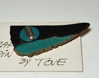 May include: A triangular ceramic pin featuring a glossy black upper section and a textured turquoise lower edge. A silver-toned metal pin backing is attached to a circular turquoise base. The pin rests on a white card with the text BY TOVE.