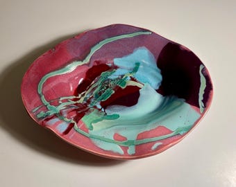 May include: An oval ceramic bowl with an irregular, wavy rim. The surface features a glossy glaze in shades of magenta, deep burgundy, and bright turquoise, with swirling, marbled patterns that blend across the interior of the dish.