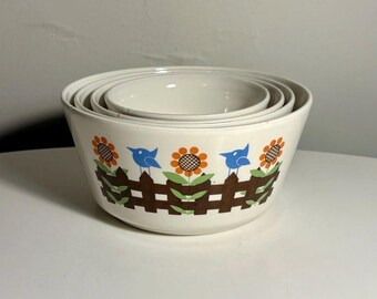 May include: A set of three nested white ceramic mixing bowls featuring a repeating pattern of brown picket fences, orange sunflowers with green leaves, and small blue birds encircling the exterior of the largest bowl in the stack.