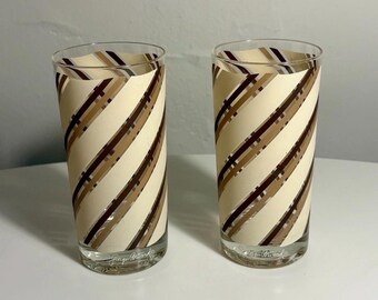 May include: Two clear glass tumblers featuring a diagonal striped pattern in shades of cream, tan, and dark brown. Each glass has a cylindrical shape and includes the signature text "Jacques Briard" printed in white near the base of the glass.