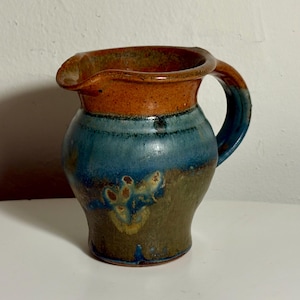 May include: A ceramic pitcher featuring a rounded body and a curved handle. The upper portion is glazed in a warm orange-brown, transitioning into a deep blue midsection, and ending in a mottled olive-green base with a wide pouring spout.