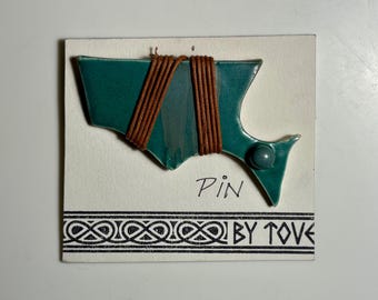 May include: A teal ceramic pin shaped like a stylised fish, wrapped with brown cord, mounted on a white card with the text Pin and By Tove, featuring a black and white decorative border pattern at the bottom of the display card.